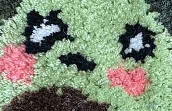 Nahaufnahme Cut Pile: Augen und Mund der Avocado mit schmalen schwarzen Konturen und rosa Wangen auf hellgrünem, plüschigem Flor.