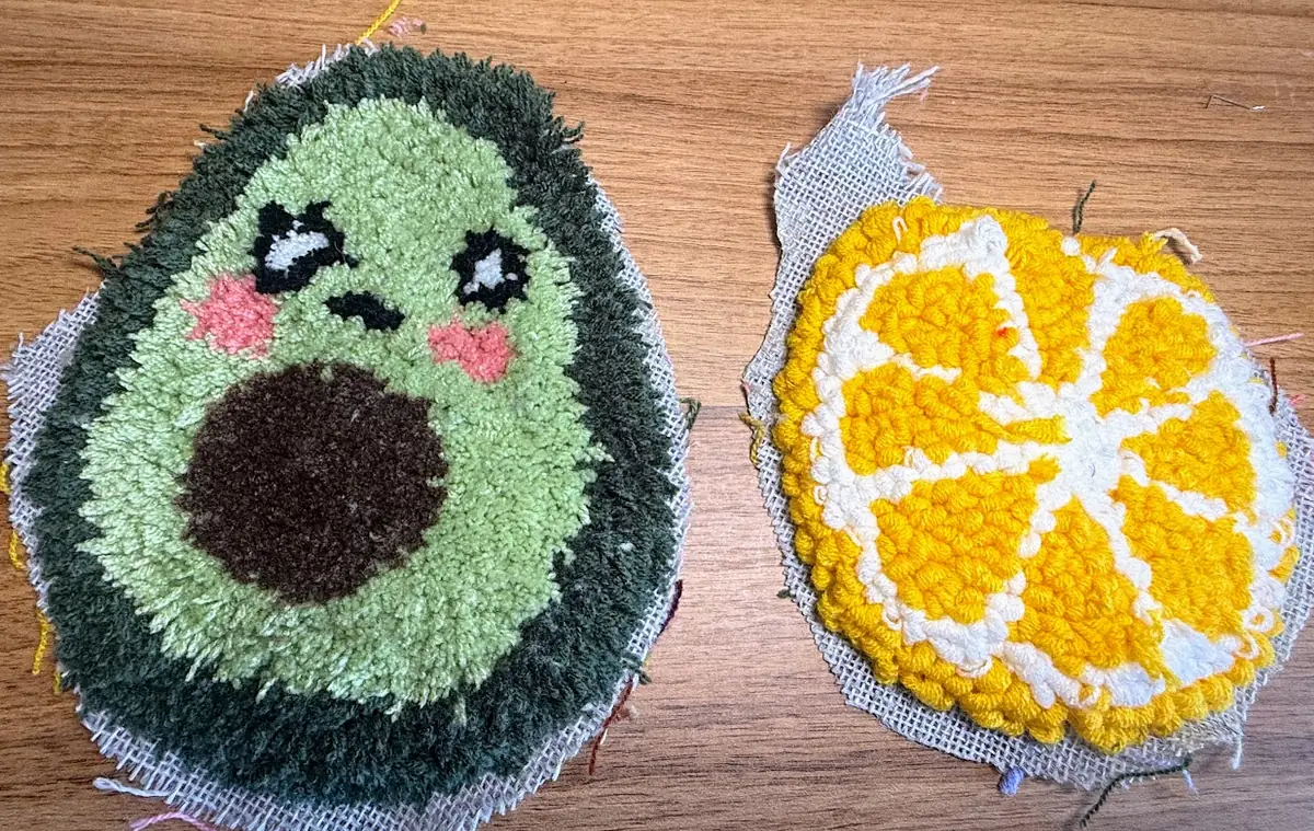 Zwei kleine getuftete Probestücke auf einem Holztisch: links eine Avocado mit Gesicht in Cut Pile mit weicher, plüschiger Oberfläche, rechts eine gelb-weiße Orangenscheibe in Loop Pile mit sichtbaren Schlingen.