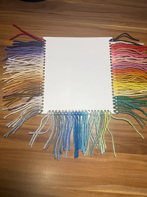 Farbkarte mit eingehängten Schurwoll-Garnproben: links Beige-, Braun-, Grau- und Lilatöne, rechts Rot-, Rosa-, Orange-, Gelb- und Grüntöne, unten verschiedene Blau- und Graublautöne.