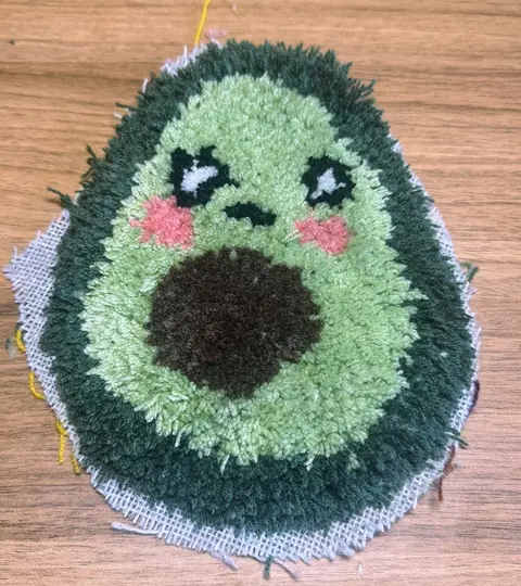 Getuftete Avocado direkt nach dem Tuften, noch auf dem Tuftingstoff: Die Konturen von Augen, Mund und Wangen wirken ausgefranst.