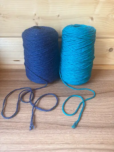 Zwei Garnkonen nebeneinander auf einem Holztisch: links dunkelblaue Schurwolle mit dickem, fest gedrehtem Faden, rechts türkises Acryl-Tuftinggarn mit dünnerem, weicherem Faden.
