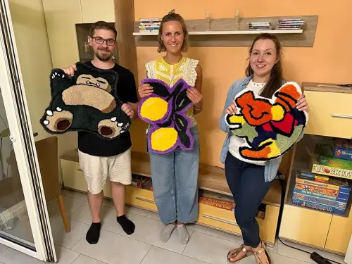 Drei Teilnehmer zeigen ihre selbstgemachten Teppiche aus dem Tufting Workshop mit bunten Motiven.